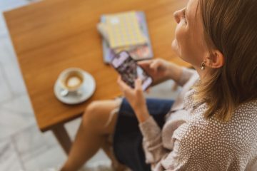 Nainen ylhäältä päin kuvattuna, istuu pöydän ääressä puhelin kädessään. Katsoo hymyillen ulos kuvasta. Pöydällä kahvikuppi ja vihkoja sekä tietokone.