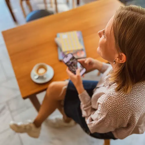 Nainen ylhäältä päin kuvattuna hymyilee ja katsoo kuvasta ulos, kädessä puhelin ja pöydällä näkyy kahvikuppi ja vihkoja.